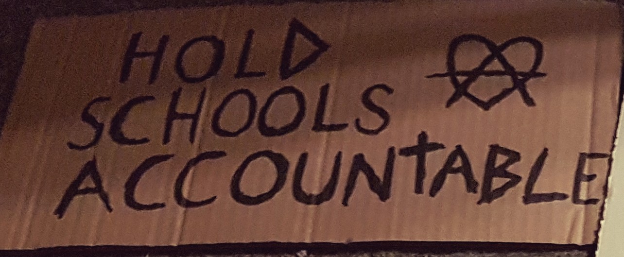 cardboard sign handpainted with the words "HOLD SCHOOLS ACCOUNTABLE" in all caps, beside an 'A' in a loveheart